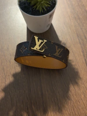 Armband  - Snyggt armband i brunt läder med klassiskt monogrammönster och guldfärgat -spänne. Insidan är ljusbrun och armbandet har en bred, stilren design som verkligen sticker ut.