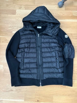 Svart dunjacka med huva från Moncler - Snygg svart dunjacka från Moncler med huva och ribbade ärmar. Jackan har två framfickor med knappar, dragkedja framtill och en Moncler-logga på ärmen. Perfekt för kalla vinterdagar och riktigt stilren look.