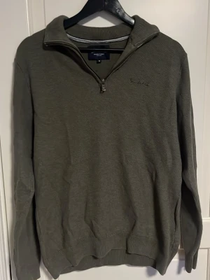 Olivgrön tröja med halv dragkedja bondelid - Olivgrön långärmad tröja med halv dragkedja och ståkrage. Tröjan har ribbade muddar vid ärmslut och nederkant samt diskret broderad logga på bröstet. Perfekt för lager på lager och enkel att matcha med jeans. 