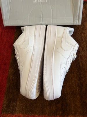 Nike Air Force 1 vita sneakers - Klassiska Nike Air Force 1 sneakers i helvitt läder med perforerad tåbox och vit sula. Skorna har den ikoniska swoosh-loggan på sidan och vita skosnören. Perfekt för dig som gillar en clean och tidlös stil.