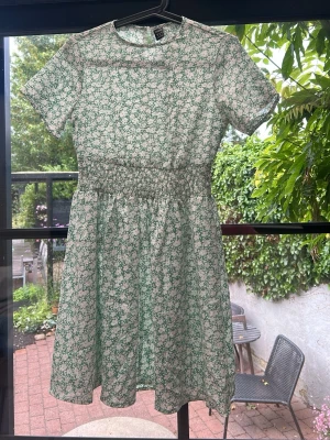 Grön blommig kortklänning - Söt grön kortklänning med vitt blommönster och smock i midjan. Klänningen har rund halsringning, korta ärmar och knapp i nacken. Perfekt för varma dagar och har en luftig passform.