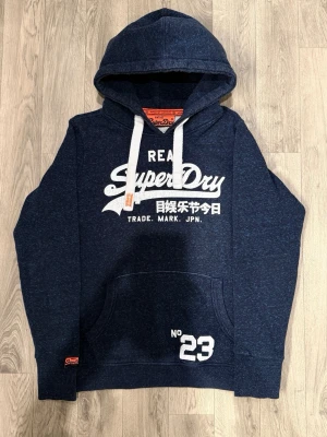 Superdry Vintage Hoodie Mörkblå Strl L (Passar M/L) - Superdry Vintage mörkblå hoodie med klassisk passform, märkt storlek L men passar bättre som normal M eller liten L. Bröstvidd 55 cm, axelbredd 42 cm, armlängd 64 cm, midja 47 cm, längd 65 cm. I fint skick.