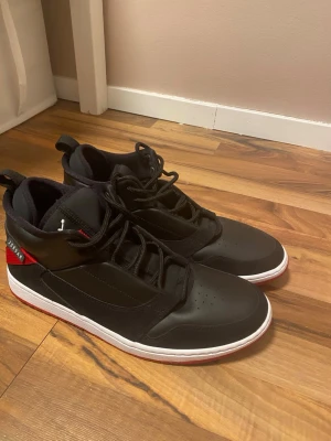 Jordan fadeaway storlek 45 - Snygga svarta Jordan sneakers med röda och vita detaljer. Skorna har högt skaft, perforerad tå och klassisk snörning. Tillverkade i syntetmaterial och textil, med en vit mellansula och röd yttersula. Perfekta för dig som gillar streetwear och sportig stil.