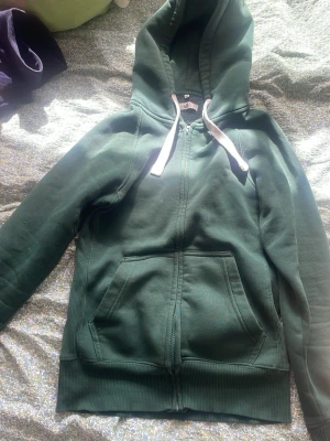 Mörkgrön hoodie med dragkedja - Mysig mörkgrön hoodie med dragkedja framtill och stora fickor. Har vit dragsko i huvan och ribbade muddar vid ärmslut och nederkant. Perfekt för chill dagar eller när du vill ha något bekvämt och enkelt.