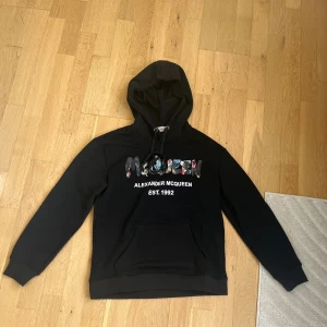 Svart hoodie Alexander McQueen - Svart hoodie från Alexander McQueen med färgglad logotyp framtill och vit text 'EST. 1992'. Tröjan har huva med snörning och en stor magficka. Perfekt för dig som gillar streetwear och vill sticka ut med ett designerplagg.