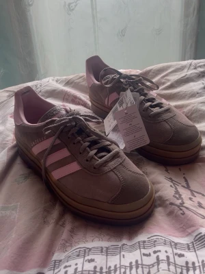Adidas Gazelle rosa och beige sneakers - Snygga Adidas Gazelle sneakers i beige mocka med ljusrosa detaljer och klassiska tre ränder på sidan. Skorna har en brun gummisula och rosa innerfoder. Perfekta för dig som vill ha en retro och trendig look.