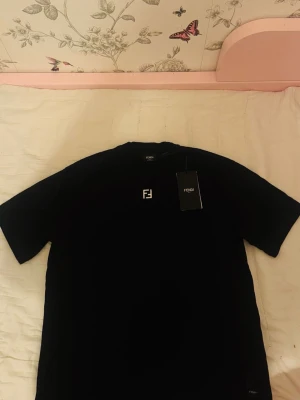 Svart t-shirt från Fendi - Snygg svart t-shirt från Fendi med klassisk logga på bröstet. Enkel och stilren design, perfekt till jeans eller shorts. T-shirten har korta ärmar och rund hals. Taggen sitter kvar.