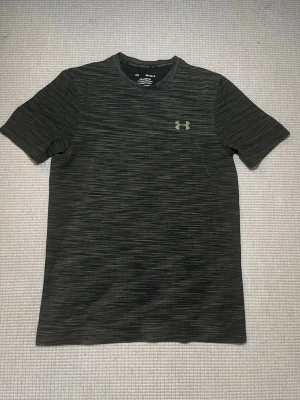 Svart tränings-tshirt Under Armour - Säljer en svart/gråmelerad tränings-tshirt från Under Armour med diskret logga på bröstet. T-shirten har rund hals, korta ärmar och är tillverkad i ett lätt och snabbtorkande material som passar perfekt till gymmet eller löpning.