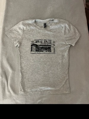 Grå t-shirt med New York tryck från HM - Snygg thigh ljusgrå t-shirt med ett svartvitt tryck av en pizzeria i Downtown New York på bröstet. T-shirten har rund hals och korta ärmar. Från divided 