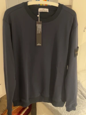 Mörkblå sweatshirt från Stone Island - Mörkblå sweatshirt från Stone Island med klassisk rund halsringning och ribbade muddar. Tröjan har den ikoniska Stone Island-loggan på vänster ärm och är tillverkad i mjuk bomull. Perfekt för en stilren och avslappnad look.