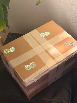 Mystery box - Säljer en brun kartonglåda som är ordentligt tejpad med vit tejp och dekorerad med flera holografiska klistermärken. Perfekt för förvaring eller frakt av olika saker.
