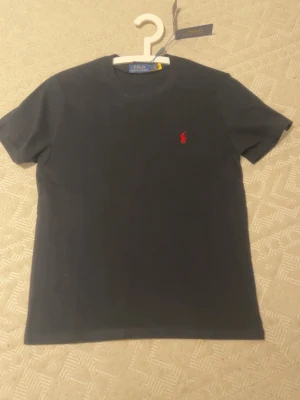 Svart t-shirt från Polo Ralph Lauren - Klassisk svart t-shirt från Polo Ralph Lauren med rund hals och korta ärmar. T-shirten har den ikoniska röda logotypen broderad på bröstet och är tillverkad i mjuk bomull för en skön känsla. Perfekt basplagg till garderoben.