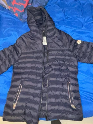 Mörkblå dunjacka med huva - Snygg mörkblå dunjacka med huva och dragkedja framtill. Jackan har två fickor med dragkedja och quiltad design. Perfekt för kalla vinterdagar och har en klassisk look som passar till det mesta.