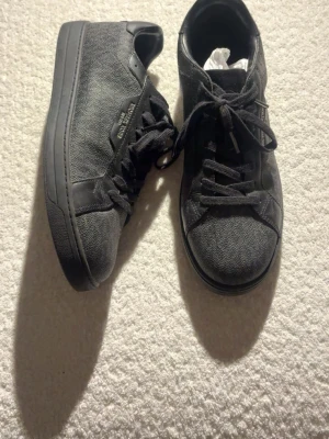 Svarta sneakers från Michael Kors - Snygga svarta sneakers från Michael Kors med diskret logomönster på sidan. Skorna har klassisk snörning och är tillverkade i en mix av syntet och skinn för en stilren look. Perfekta för dig som vill ha en clean och modern stil.