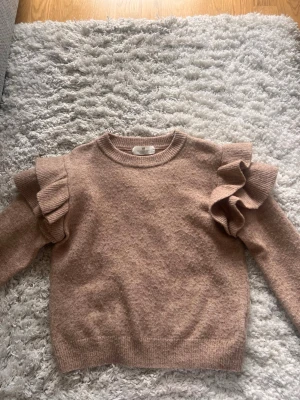 Beige stickad tröja med volang - Supersöt beige stickad tröja med volangdetaljer på axlarna. Tröjan har rund halsringning, långa ärmar och ribbade muddar. Perfekt för dig som gillar en trendig och feminin look.