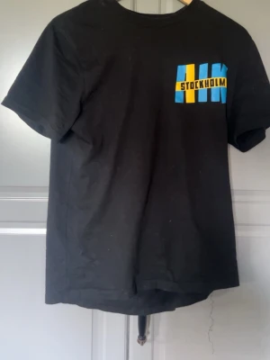 Svart t-shirt med Sverige-tryck - Cool svart t-shirt med tryck av 'STOCKHOLM' i blått och gult framtill och stort 'SVERIGE' i vitt och gult på ryggen. Klassisk passform och rund hals. Perfekt för dig som vill visa lite svensk stolthet.