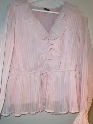 Ljusrosa blus med volang från Vero Moda - Söt ljusrosa blus från Vero Moda med långa ärmar, volangdetaljer längs ringningen och omlottkänsla. Blusen har knyt i midjan och är tillverkad i ett lätt, plisserat material som ger en luftig känsla. Storlek L, aldrig använd endast tvättad. 