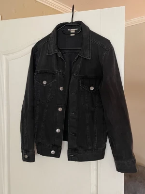 Svart jeansjacka - Snygg urtvättad svart jeansjacka med silverfärgade knappar och två bröstfickor. Är storlek 36, men är oversized. Perfekt att slänga över en hoodie eller t-shirt för en cool och avslappnad look.