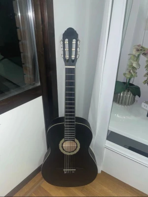 Svart klassisk akustisk gitarr - Säljer en svart klassisk akustisk gitarr med sex strängar och dekorativ rosett runt ljudhålet. Gitarren har vita detaljer längs kanterna och huvudet, samt silverfärgade stämskruvar. Perfekt för dig som vill börja spela eller bara jamma hemma.
