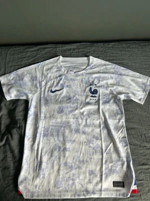 Vit Frankrike matchtröja Nike - Säljer en vit matchtröja från Nike med Frankrikes landslagsmärke på bröstet. Tröjan har ett diskret blått mönster över hela tyget, korta ärmar och rund hals. Perfekt för dig som älskar fotboll och vill stötta Les Bleus på matchdag.