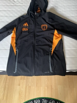 Fotbolls tracksuit  - Snygg svart sportjacka med orange detaljer och huva. Jackan har dragkedja framtill, två fickor med reflexdetaljer och tryck på bröstet, inklusive Paris FC-logga. Perfekt för träning eller vardagsbruk. Tillverkad i lätt och funktionellt material.