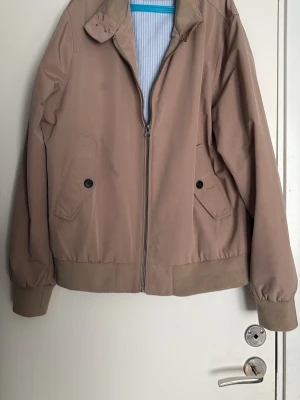 Beige bomberjacka från Piombo - Snygg beige bomberjacka från Piombo med dragkedja framtill, ribbade muddar och krage i kontrastmaterial. Två fickor med knappstängning och diskret randigt innerfoder. Perfekt för dig som vill ha en stilren och enkel jacka.
