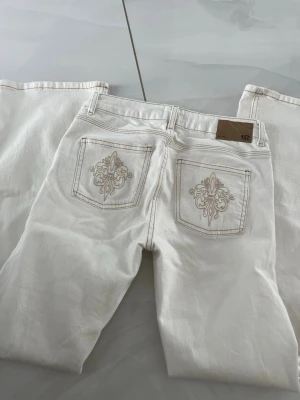 Vita jeans med broderade bakfickor - Säljer ett par vita jeans med snygga broderade detaljer på bakfickorna. Jeansen har utsvängda ben, klassisk femficksmodell och bälteshällor. Materialet är kraftigt denim i bomull och de har en stilren look som passar till det mesta. Oanvända.