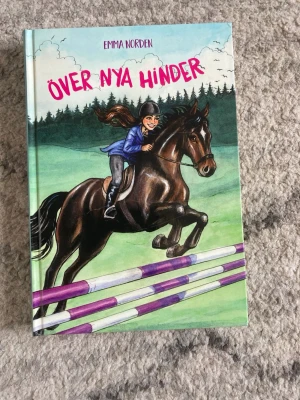 Över nya hinder av Emma Norden - En färgglad bok med hårda pärmar, illustrerad med en tjej som hoppar hinder på en brun häst. Omslaget har gröna och blå toner med lila och vita hinderstavar. Perfekt för dig som gillar hästar och äventyr.