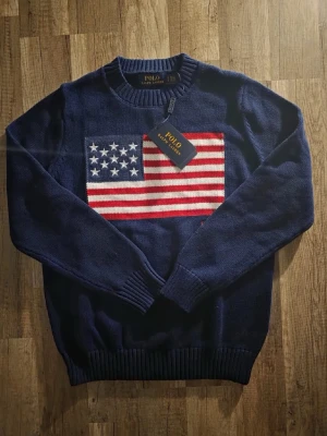 Mörkblå stickad tröja Polo Ralph Lauren - Snygg mörkblå stickad tröja från Polo Ralph Lauren med amerikansk flagga på bröstet. Tröjan har rund halsringning, ribbade muddar och nederkant. Perfekt för dig som vill ha en klassisk och trendig look.