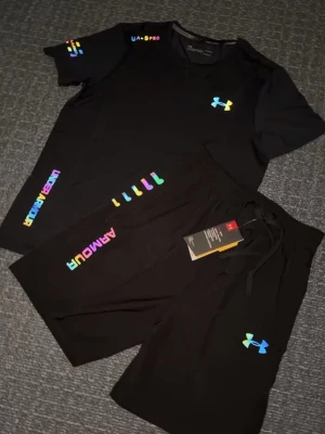 Under Armour svarta summer set - Svart träningsset med t-shirt och shorts från Under Armour. Båda plaggen har färgglada loggor och detaljer i regnbågsfärger. Perfekt för träning och sportiga aktiviteter. Materialet känns lätt och andas bra.