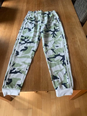 Camo mjukisbyxor från Adidas - Säljer ett par mjukisbyxor från Adidas med grön, vit och svart camomönster. Byxorna har resår i midjan med dragsko, vita muddar vid bensluten och klassiska Adidas-ränder längs sidorna. Perfekta för en chill och sportig look.