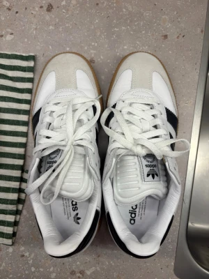 Adidas vita sneakers med svarta ränder - Säljer ett par klassiska Adidas sneakers i vitt läder med svarta ränder och beige mockadetaljer framtill. Skorna har en brun gummisula och vit plös med Adidas-logga. Perfekta för dig som gillar retrostil och streetwear.