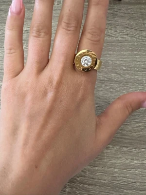 Guldfärgad ring med stor kristall från LenMer - Säljer en chunky guldfärgad ring med en stor, glittrande kristall i mitten. Ringen har en bred design och text runt stenen som ger en cool och modern vibe. Perfekt för dig som gillar statement-smycken och vill sticka ut. Ringen är i storlek 7 och helt oanvänd! OBS: FRAKTAR INOM 24H!🩷