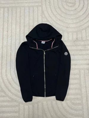 Svart hoodie från Moncler med dragkedja - Svart hoodie från Moncler med dragkedja framtill och klassisk huva. Tricolore-detalj på insidan av kragen och Moncler-logga på vänster ärm. Mjuk bomullskänsla och två fickor framtill. Perfekt för en stilren och avslappnad look.