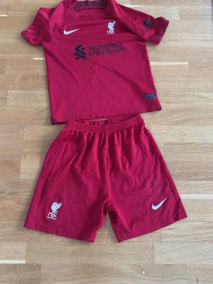 Liverpool matchställ röd Nike - Säljer ett rött Liverpool matchställ från Nike med klubbmärke och sponsortryck på tröjan samt shorts. Perfekt för fotbollsträning eller att bära när du hejar på ditt favoritlag. Materialet är lätt och andas bra.