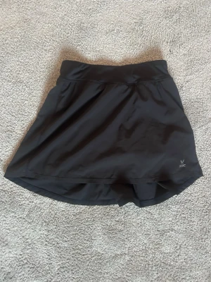 Svart sportig skort från SOC - Snygg svart skort från SOC med bred midjeresår och inbyggda shorts under kjolen. Tillverkad i ett stretchigt och snabbtorkande material som passar perfekt för träning eller aktiva dagar. Enkel och stilren design med liten logga på sidan.