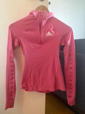 Rosa träningströja från Adidas - Snygg rosa långärmad träningströja från Adidas med halv dragkedja framtill och vit logga på bröstet. Tillverkad i ett stretchigt funktionsmaterial som passar perfekt till löpning eller gym. Figurnära passform och hög krage för extra komfort.