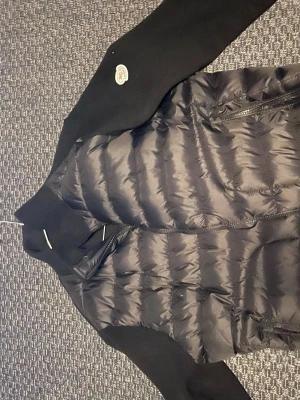 Svart dunjacka från Moncler - Snygg svart dunjacka från Moncler med quiltad kropp och stickade ärmar. Jackan har dragkedja framtill, två sidofickor med dragkedja och en liten Moncler-logga på ärmen. Perfekt för kalla vinterdagar och riktigt trendig look.