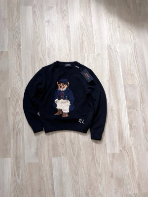 Ralph lauren björn stickad - Säljer en mörkblå stickad tröja med ett stort broderat motiv av en björn på framsidan. Björnen har blå jacka, beige byxor och mössa. Tröjan har rund halsringning och ribbade muddar. Perfekt för dig som gillar unika och lekfulla plagg.