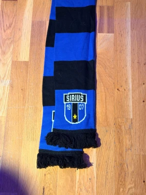 Sirius halsduk  - HELT NY OCH OANVÄND SIRIUS HALSDUK KÖPTE FÖR 250kr SÅ PASSA PÅ ATT KÖP FÖR ETT OTROLIGT PRISY? Sublimerad halsduk i polyester, breda blåsvarta ränder och emblemet i ändarna.
