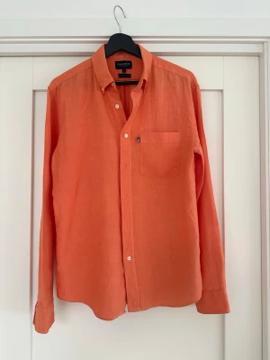Orange linneskjorta lexington  - Säljer en snygg orange skjorta i linne med klassisk krage och bröstficka. Skjortan är i mycket bra skick och har endast använts fåtalet gånger. Det enda defekt som finns är de svarta sträcken på armen och baktill (se sista 3 bilderna). Dock inget som märks vid användning. Nypriset 1199 mitt 249kr.