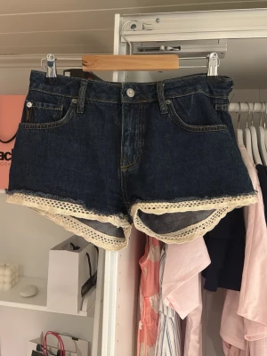 Blå jeansshorts med spets från Superdry - Säljer ett par mörkblå jeansshorts från Superdry med snygg spetsdetalj längs bensluten. Shortsen är gjorda i ekologisk bomull och har klassisk femficksdesign, knappgylf och midwaist passform. Perfekta för sommaren!