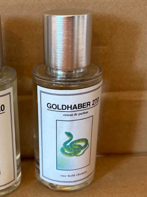Goldhaber 270 parfymflaska - Stilren parfymflaska med metallock och transparent glas. Etiketten har en grön orm och texten 'GOLDHABER 270' samt 'extrait de parfum'. Flaskan har en modern och minimalistisk design som passar perfekt på badrumshyllan.