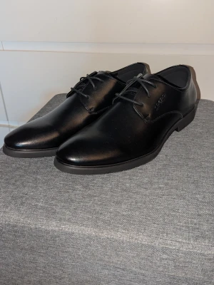Svarta klassiska loafers i skinn - Säljer ett par stilrena svarta loafers i slätt skinn med rundad tå och diskreta sömmar. Skorna har en låg klack och svarta snören för en clean look. Perfekta för dig som gillar enkel och tidlös stil.