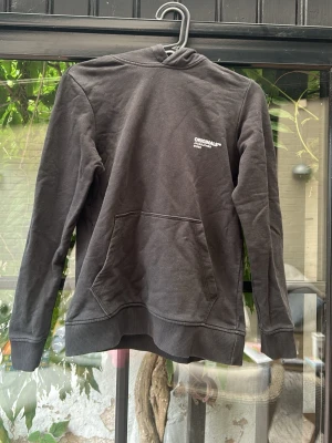 Svart hoodie från Jack & Jones - Säljer en svart hoodie från Jack & Jones Originals med vit logga på bröstet. Hoodien har huva och en stor magficka framtill. Tillverkad i mjukt bomullsmaterial och har ribbade muddar vid ärmar och nederkant. Perfekt för en avslappnad stil. Har ett litet hål där bak nere vid