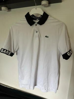 Vit Lacoste piké med GAME-detalj - Vit pikétröja från Lacoste med svart krage och svarta muddar på ärmarna där det står 'GAME' i stora vita bokstäver. Klassisk Lacoste-logga på bröstet. Tillverkad i bomull och har korta ärmar samt knappar vid halsen.