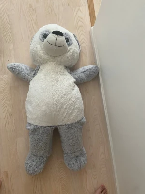 Stor grå och vit nallebjörn - Mysig och stor nallebjörn i mjuk plysch med grå kropp, vita detaljer på mage och ansikte samt mörkare nos. Perfekt som gosedjur eller dekoration i sovrummet. Nallen har utsträckta armar och ben och ett vänligt ansikte.