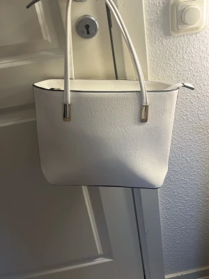 Vit handväska med silverdetaljer - Stilren vit handväska i konstläder med två långa axelband och silverfärgade metalldetaljer. Väskan har en enkel design med dragkedja upptill och en praktisk ytterficka med dragkedja på baksidan. Perfekt för dig som gillar minimalistisk och modern stil.