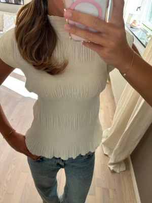 Vit ribbad topp med kort ärm - Säljer en söt vit topp med ribbad struktur och korta ärmar. Toppen har en figurnära passform och lätt vågig kant nedtill. Perfekt till jeans eller kjol för en fräsch look.