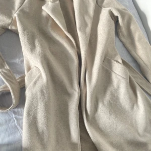 Beige kappa från Vero Moda - Säljer en stilren beige kappa från Vero Moda med bälte i midjan och klassisk krage. Jackan har långa ärmar och två fickor framtill. Perfekt för dig som vill ha en tidlös och enkel look.
